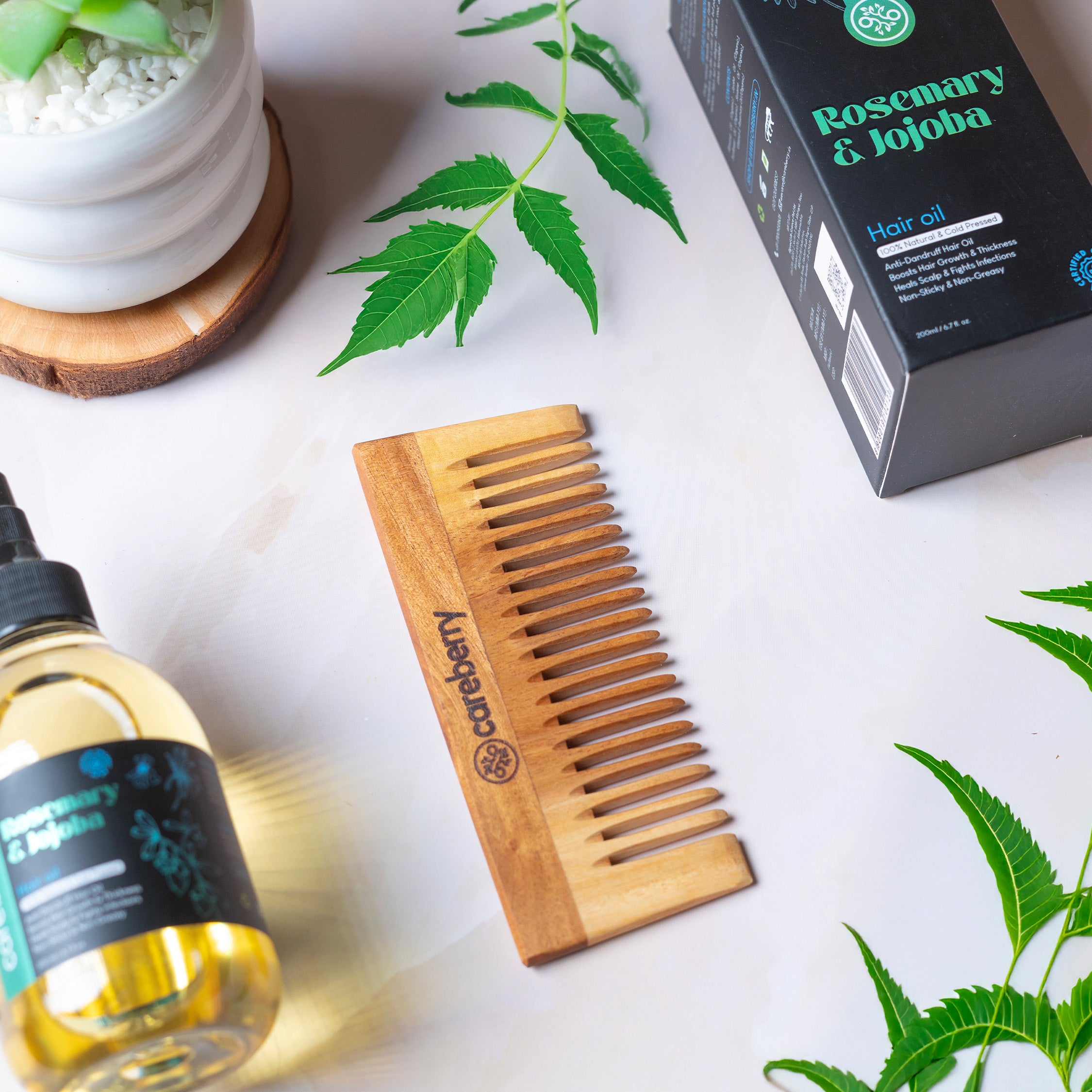Careberry wooden neem comb displayed in flat lay with rosemary jojoba hair oil bottle, black branded box, fresh green neem leaves and natural herbs on white background.